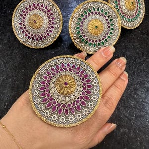 May include: Ornate, large statement rings with a silver and gold-tone design. The rings feature a central design with a circular pattern of red or green gemstones and clear crystals. One ring is on a hand.