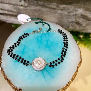 May include: A silver bracelet with a white flower charm and black beads. The bracelet is on a blue and gold background.