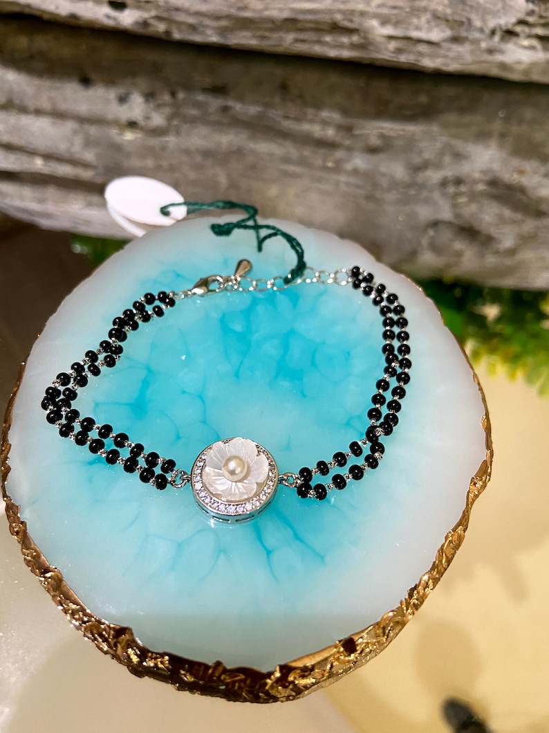 May include: A silver bracelet with a white flower charm and black beads. The bracelet is on a blue and gold background.