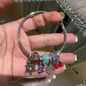 May include: A silver bracelet with various charms, including a snowflake, a purple flower, a clear flower, and red cherries. The charms are adorned with crystals and colorful accents. The bracelet is held in a hand.