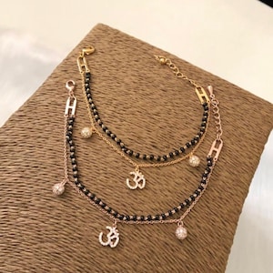 May include: A gold chain bracelet with black beads and three charms. The charms are a small ball with rhinestones, and two Om symbols with rhinestones.