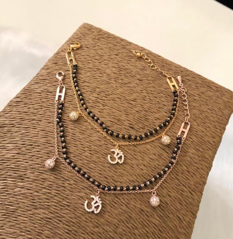 May include: A gold chain bracelet with black beads and three charms. The charms are a small ball with rhinestones, and two Om symbols with rhinestones.