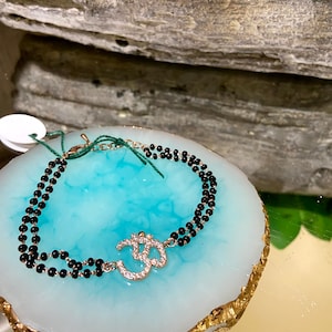 May include: A black beaded bracelet with a gold Om charm adorned with sparkling rhinestones.