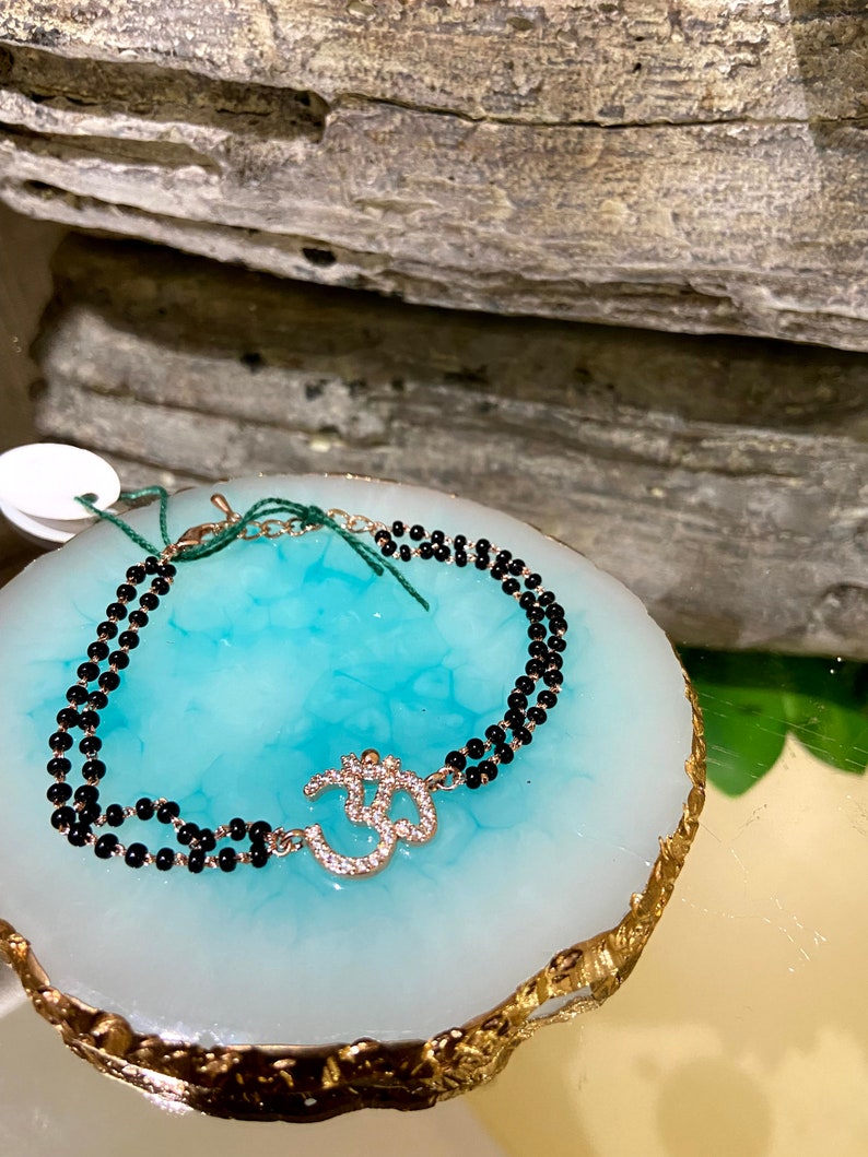 May include: A black beaded bracelet with a gold Om charm adorned with sparkling rhinestones.