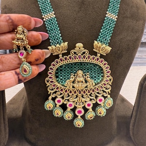May include: An ornate gold-toned necklace and earring set. The necklace features a large pendant with a central oval design filled with green beads, surrounded by red gemstones. The necklace has a strand of green beads. The earrings have a matching design.