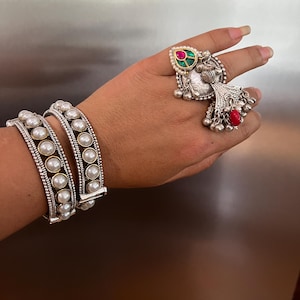 May include: A hand displays two pearl-studded bracelets and a silver ring with a red and green gemstone. The bracelets have a silver band with a row of pearls. The ring features a teardrop-shaped green and pink stone and dangling silver bells.