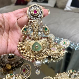 May include: A pair of gold earrings with green and red stones, pearls, and intricate detailing. The earrings are in the shape of a crescent moon with a floral design.