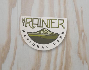 May include: A semi-circular sticker featuring a graphic illustration of Mt. Rainier. The design includes the text MT RAINIER NATIONAL PARK in dark green lettering, with a mountain peak in white, olive green, and dark brown colors on a white background.