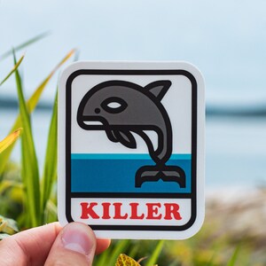 May include: A white sticker with a black outline features a cartoon killer whale jumping out of blue water. The text "KILLER" is printed in red letters below the whale.