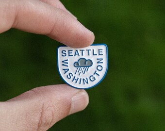 Puede incluir: Un pin de esmalte blanco y azul con el texto "SEATTLE WASHINGTON" y una nube de lluvia azul con gotas de lluvia.