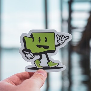 May include: A green cartoon sticker of the state of Washington with arms and legs. The sticker is shaped like the state and has a friendly face with a smile. The sticker is holding its hand up in a 'hang loose' gesture.