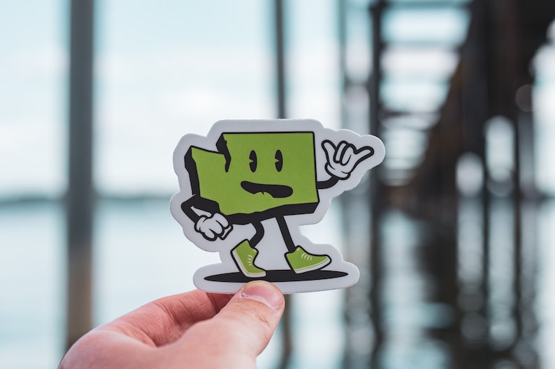 May include: A green cartoon sticker of the state of Washington with arms and legs. The sticker is shaped like the state and has a friendly face with a smile. The sticker is holding its hand up in a 'hang loose' gesture.