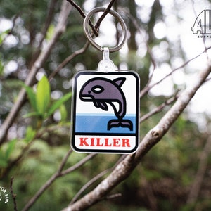May include: A white and blue key chain with a black and gray cartoon killer whale jumping out of the water. The key chain has the word "KILLER" printed in red letters.