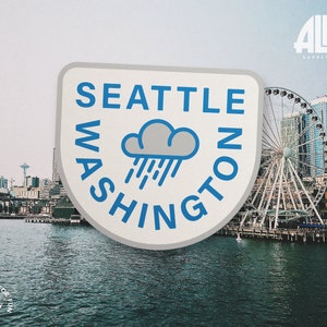 May include: A white and blue sticker with the text "Seattle Washington" in a circle around a gray cloud with blue rain drops. The sticker is in front of a view of the Seattle skyline with the Ferris wheel.