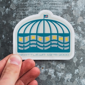 May include: A white sticker featuring a blue and grey illustration of the Kingdome, a domed stadium in Seattle, Washington. The sticker includes the text "KINGDOME" in yellow lettering and "SEATTLE, WA 1976-2000" in black lettering.