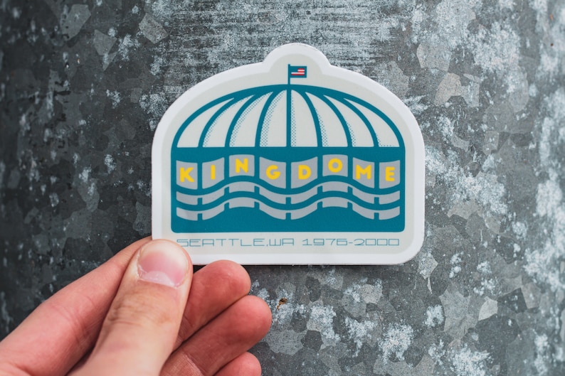 May include: A white sticker featuring a blue and grey illustration of the Kingdome, a domed stadium in Seattle, Washington. The sticker includes the text "KINGDOME" in yellow lettering and "SEATTLE, WA 1976-2000" in black lettering.