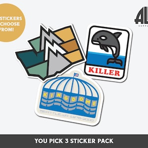 May include: A sticker pack featuring three designs: a blue and white Seattle Kingdome sticker, a blue and white orca whale sticker with the word "Killer" in red, and a green, blue, and brown mountain range sticker. The stickers are part of a "You Pick 3 Sticker Pack" and are from the company "Alki Supply Co."
