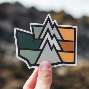 May include: A sticker with a geometric design of mountains in shades of green, grey, orange, and brown. The sticker is shaped like a state outline.