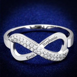 May include: A silver infinity ring with a row of small clear stones on each side. The ring is engraved with the names "Stephanie" and "Kevin".