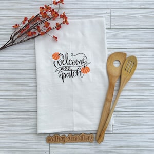 Puede incluir: Un paño de cocina blanco con texto bordado en negro que dice welcome to our patch junto a dos calabazas naranjas. El paño se acompaña de dos utensilios de cocina de bambú marrón claro y una rama decorativa con pequeñas bayas naranjas.