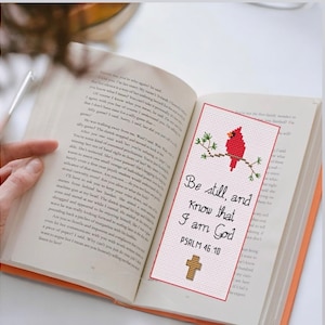 May include: A red cardinal bird sits on a branch with green leaves. The bookmark is white with red and gold accents. The text reads "Be still, and know that I am God. Psalm 46:10".
