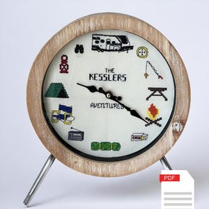 Puede incluir: Un reloj de madera con una esfera blanca y agujas negras. La esfera del reloj está decorada con un diseño de punto de cruz que presenta iconos relacionados con el camping y las autocaravanas. El texto "The Kessler's Adventures" está bordado en el centro de la esfera del reloj.