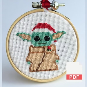Puede incluir: Un bordado de punto de cruz de un personaje alienígena verde con un gorro de Papá Noel rojo y un abrigo marrón, en un aro de madera. El diseño está sobre tela blanca, con una cinta roja en la parte superior. Un icono PDF está en la esquina inferior derecha.