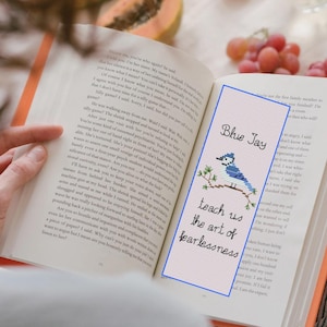 Puede incluir: Un pájaro azul arrendajo con un pecho blanco y marcas negras está posado en una rama con hojas verdes. El marcapáginas es rosa con las palabras "Blue Jay" y "teach us the art of fearlessness" en texto negro.