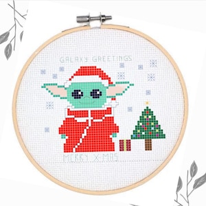 Puede incluir: Un bastidor de bordado circular de madera con un patrón de punto de cruz de una criatura verde con gorro y abrigo de Papá Noel rojos. El texto GALAXY GREETINGS y MERRY X-MAS está bordado en azul, junto a un pequeño árbol de Navidad.
