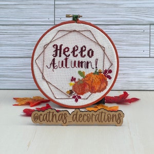 Puede incluir: Un bastidor de bordado redondo con tela blanca y el texto Hello Autumn bordado en rojo oscuro, decorado con calabazas naranjas y hojas pequeñas. Un letrero de madera al frente muestra el texto @cathas_decorations.