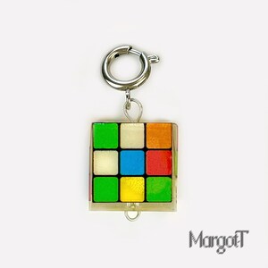 以下が含まれることがあります： シルバーの留め金が付いた、カラフルなルービックキューブのチャーム。 キューブは、赤、オレンジ、黄色、緑、青、白の正方形を持つ、クラシックな3x3デザインです。 チャームはシルバーの留め金に取り付けられています。 右下には「MargotT」の文字があります。