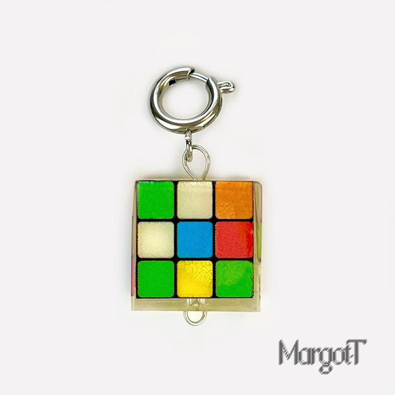 以下が含まれることがあります： シルバーの留め金が付いた、カラフルなルービックキューブのチャーム。 キューブは、赤、オレンジ、黄色、緑、青、白の正方形を持つ、クラシックな3x3デザインです。 チャームはシルバーの留め金に取り付けられています。 右下には「MargotT」の文字があります。