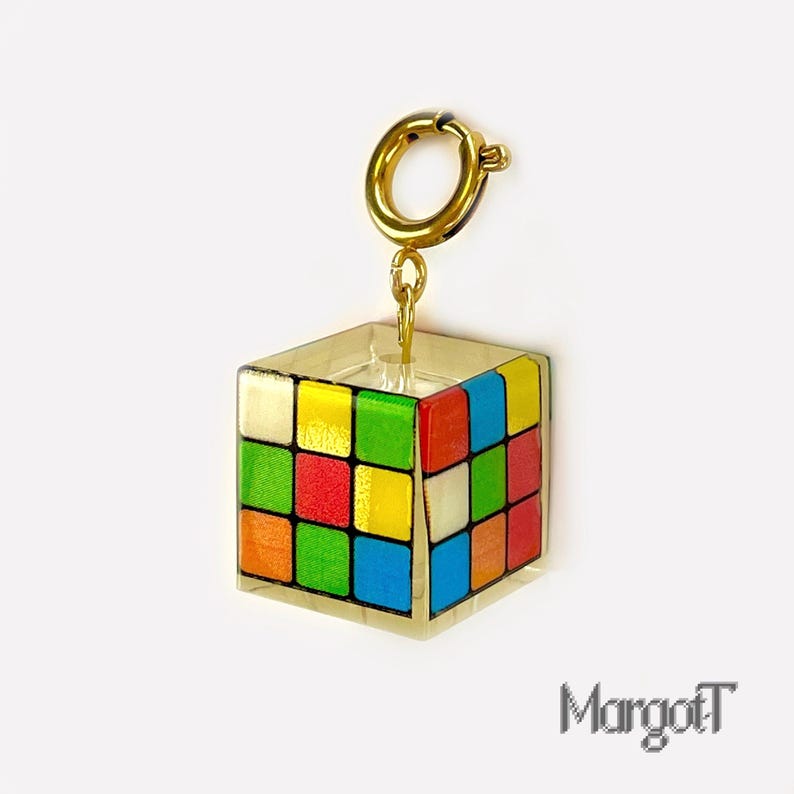 以下が含まれることがあります： カラフルなルービックキューブのデザインが施された、透明な立方体型のチャーム。チャームはゴールドカラーの留め金とチェーンから吊り下げられています。チャームにはゴールドカラーの縁取りがあります。テキスト「MargotT」が右下にあります。