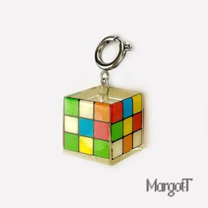 以下が含まれることがあります： カラフルなルービックキューブのチャーム、シルバーカラーの留め金付き。キューブは赤、青、緑、オレンジ、黄色、白の正方形で構成されています。チャームはシルバーのリングから吊り下げられています。「MargotT」という名前が右下に印刷されています。