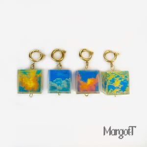 Puede incluir: Cuatro colgantes cuadrados con cierres dorados. Cada colgante presenta un diseño diferente con temática de cielo: un atardecer amarillo y naranja, un cielo azul con nubes blancas, un atardecer con tonos rosas y naranjas, y un cielo nublado amarillo y azul. Sobre fondo blanco.