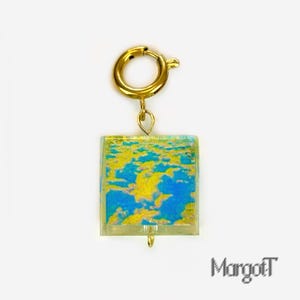 Puede incluir: Un cierre dorado con un colgante cuadrado. El colgante presenta un diseño abstracto azul y amarillo. La joya es probablemente un pendiente o un charm. El nombre del artista, MargotT, está en la esquina inferior derecha.