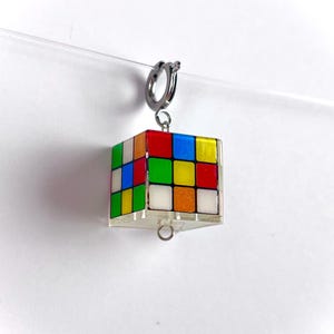以下が含まれることがあります： シルバーカラーの留め金が付いた、カラフルなルービックキューブのチャーム。赤、オレンジ、黄色、緑、青、白の正方形が特徴です。細い透明な糸で吊り下げられており、浮遊感があります。