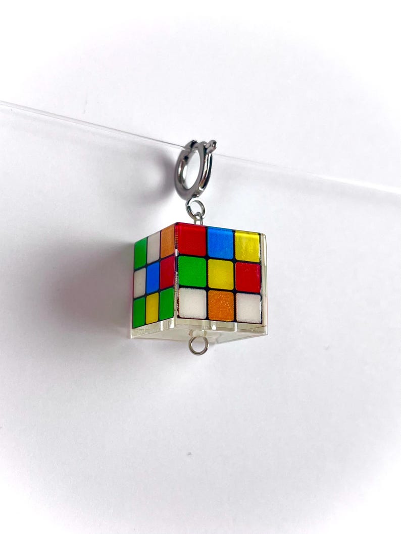 以下が含まれることがあります： シルバーカラーの留め金が付いた、カラフルなルービックキューブのチャーム。赤、オレンジ、黄色、緑、青、白の正方形が特徴です。細い透明な糸で吊り下げられており、浮遊感があります。