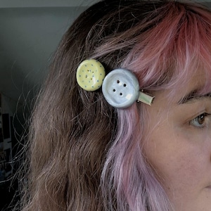 Pode incluir: Dois ganchos de cabelo com ornamentos circulares em forma de botão. Um tem um botão amarelo brilhante com pequenos pontos azuis, enquanto o outro é um botão cinzento claro maior, liso, com quatro orifícios centrais numa base de metal.