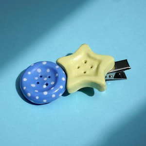 Pode incluir: Um gancho de cabelo com um botão azul redondo com bolinhas brancas ao lado de um botão amarelo pálido em forma de estrela. O gancho é feito de metal prateado e está preso à parte de trás do botão em forma de estrela.