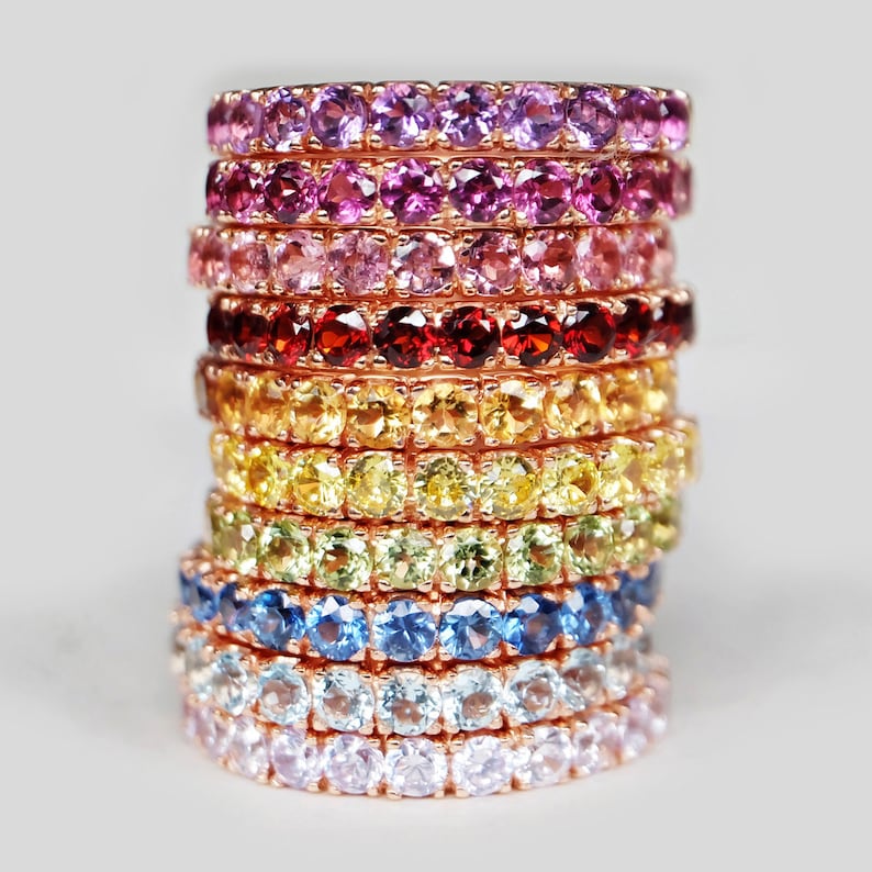May include: A stack of colourful gemstone rings in a rainbow pattern. The rings are made of rose gold and feature a variety of gemstones, including amethyst, pink sapphire, ruby, citrine, peridot, blue topaz, aquamarine, and rose quartz.