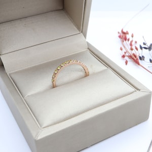 May include: A delicate gold band ring with a row of small, yellow gemstones. The ring is displayed in a beige ring box.