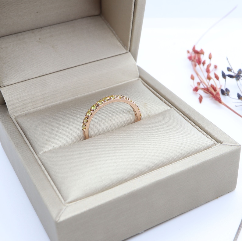 May include: A delicate gold band ring with a row of small, yellow gemstones. The ring is displayed in a beige ring box.