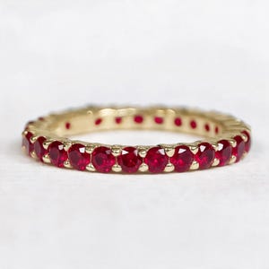 May include: An eternity band ring featuring a continuous row of round-cut, vibrant red rubies set in a polished yellow gold metal band. The circular design showcases multiple faceted gemstones held securely by small gold prongs in a classic setting.