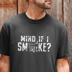 May include: Dark gray t-shirt with the text "MIND IF I SMOKE?" in white, along with a graphic of a smoker. The shirt is short-sleeved and made of a soft material. The design is centered on the chest.
