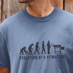 May include: Blue t-shirt with the text "EVOLUTION OF A PITMASTER" and a graphic depicting the evolution of man to a person grilling on a barbecue. The shirt is a solid blue color.