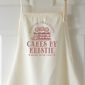 May include: White apron with a pink design that says "Custom Creations Cakes by Kristie Made with Love".