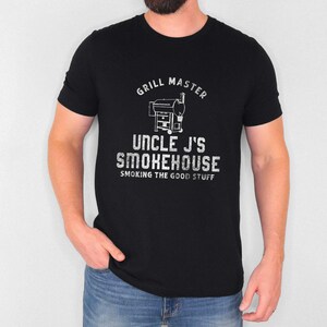 May include: Black t-shirt with a white graphic that reads "GRILL MASTER" above an illustration of a smoker. Below the smoker, the text reads "UNCLE J'S SMOKEHOUSE" and "SMOKING THE GOOD STUFF".