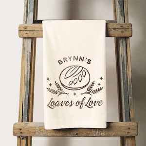May include: A white kitchen towel with a brown graphic of a loaf of bread and the text "Brynn's Loaves of Love".