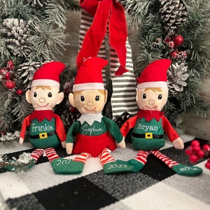May include: Three plush Christmas elves with personalized names, Frankie, Sophia, and Bryan, sitting on a black and white checkered surface. Each elf is wearing a red hat, green pants, and a red and white striped shirt. The elves are holding a sign with the year 2023 on it.